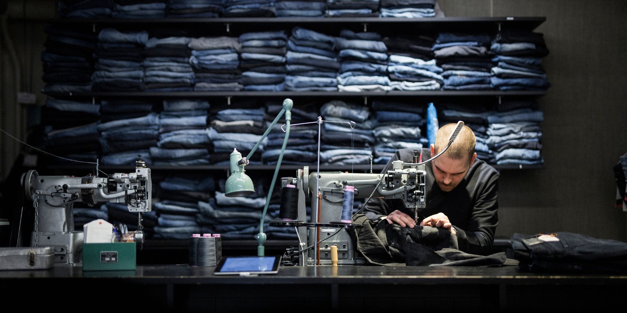 man mending Nudie Jeans with pile of jeans for repair in the background