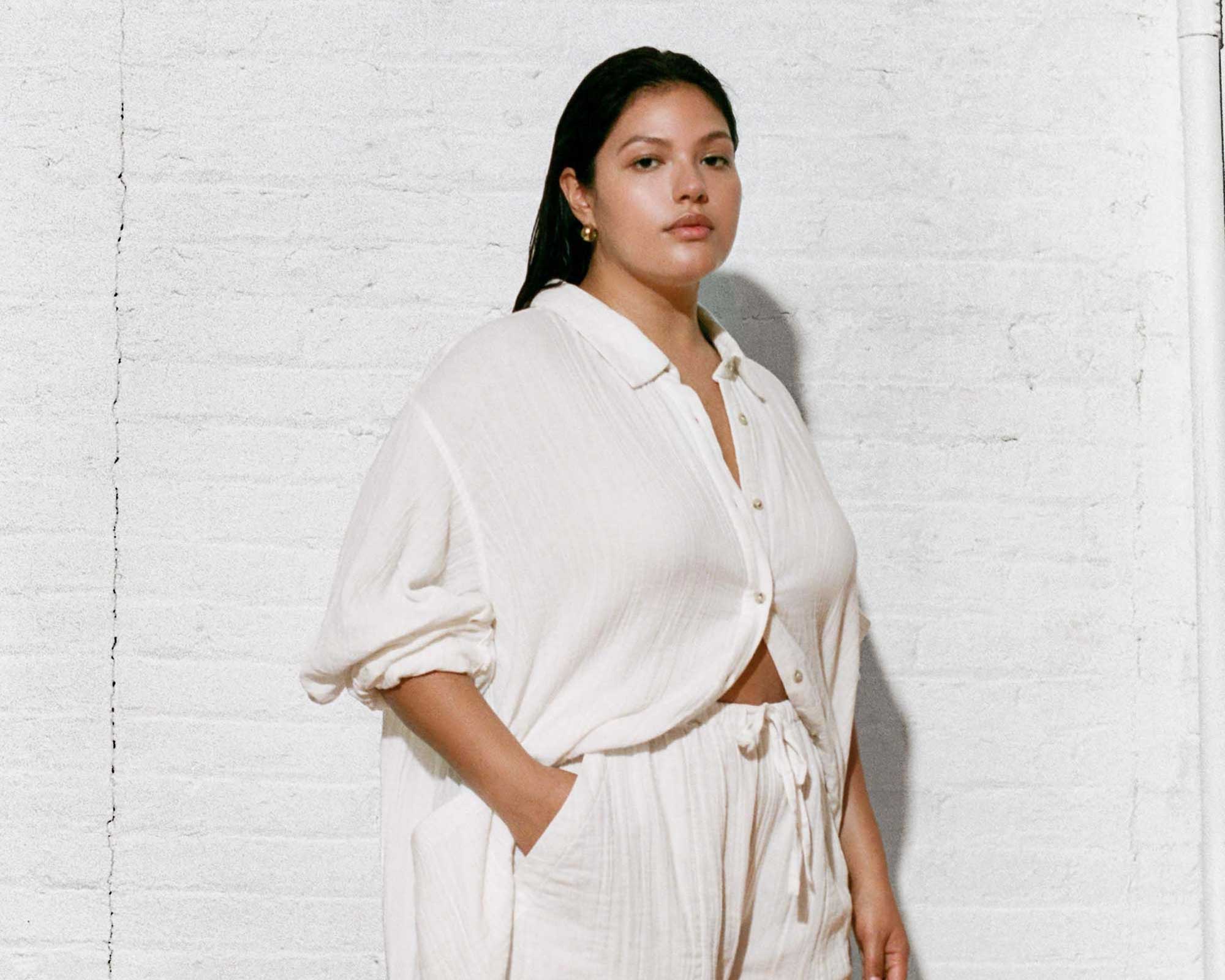 Plus size person in a cream linen button up and pants by FRANC standing with their hand in their pocket against a white brick background.