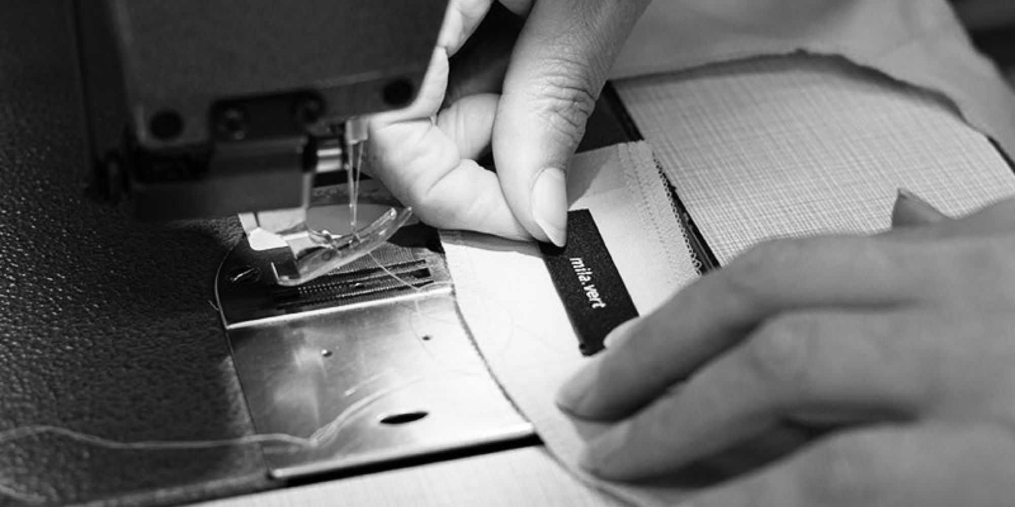 A close up of someone sewing on a Mila.Vert label to a garment, stylised in black and white.