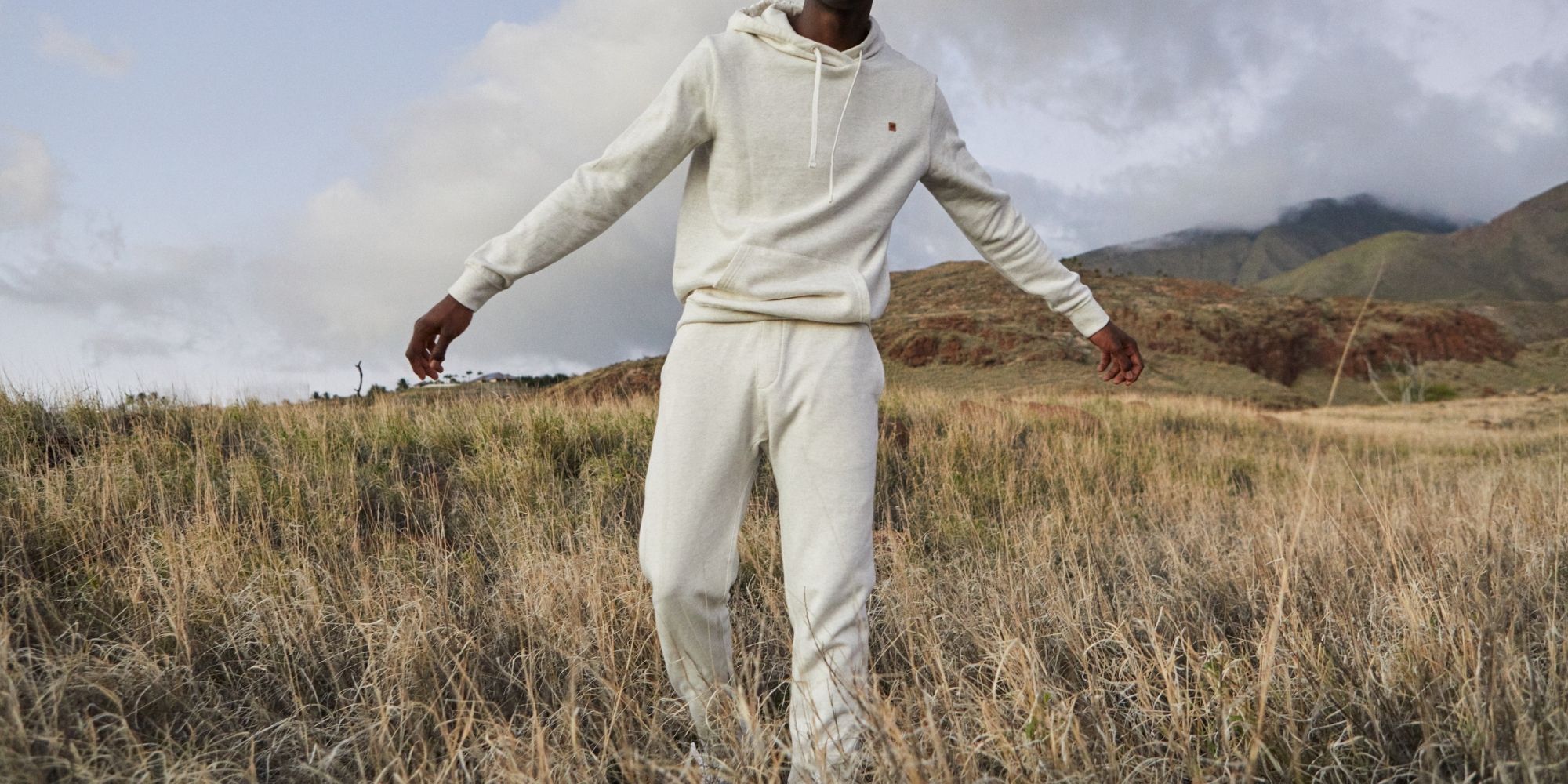 Someone standing outdoors in a field near mountains wearing a white sweatsuit set by tentree.
