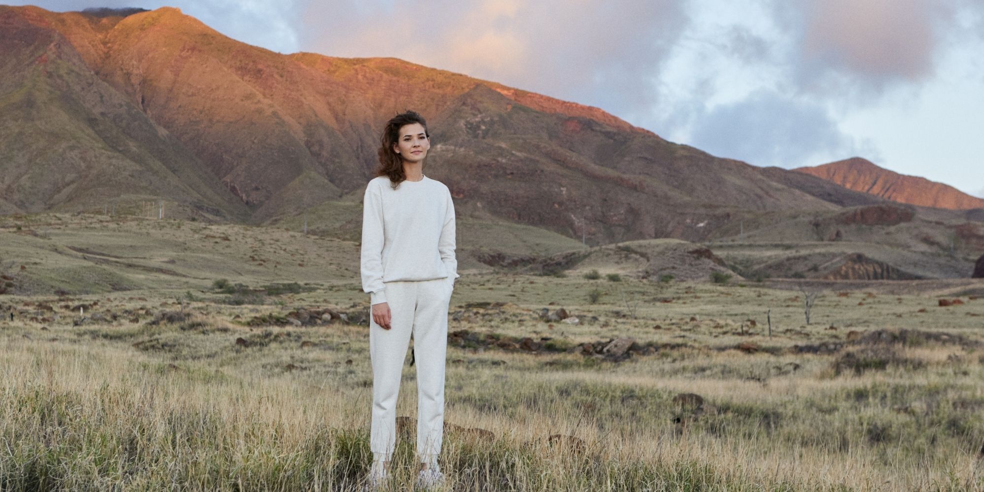 Someone standing outdoors in a field near mountains wearing a white sweatsuit set by tentree.