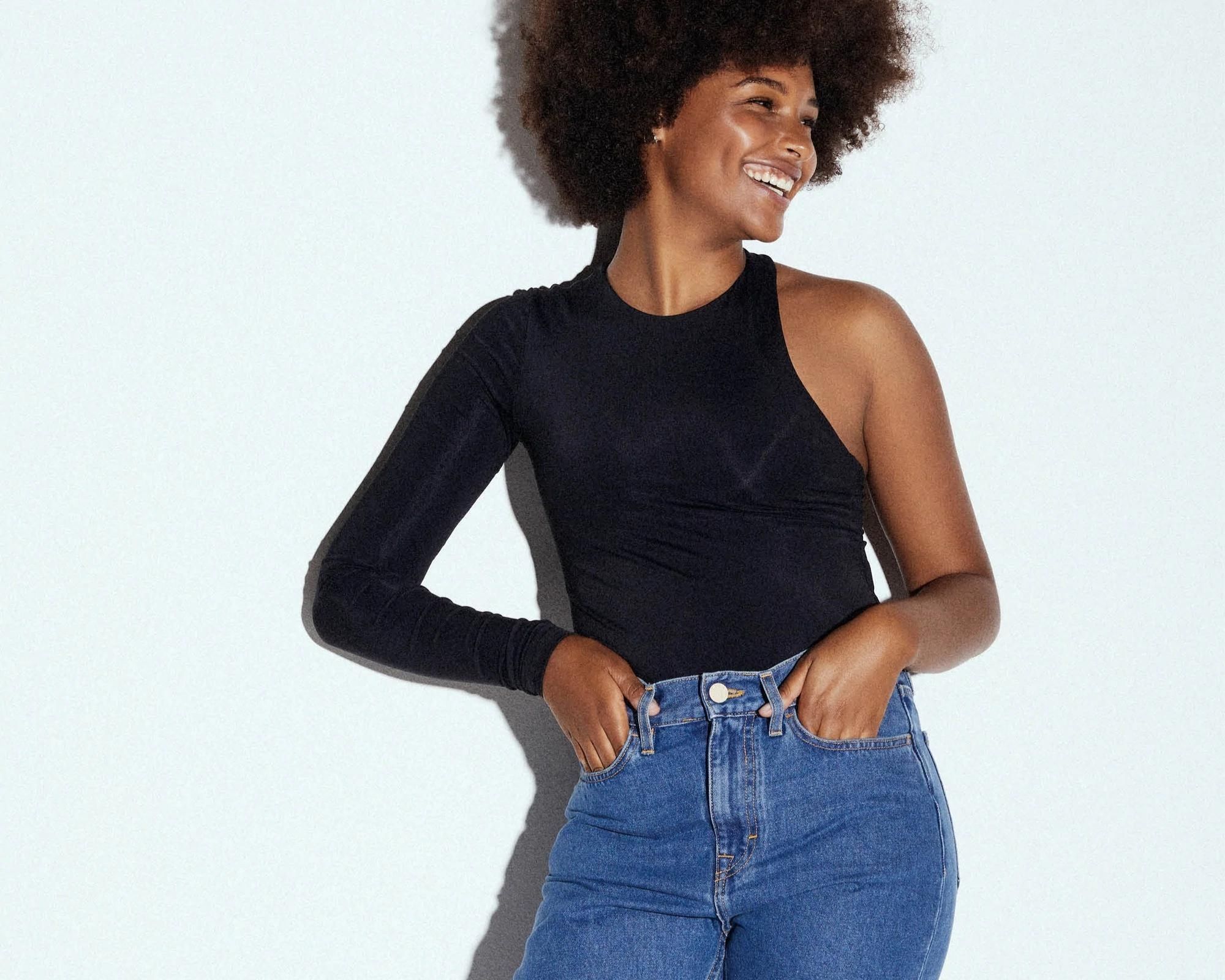 A model smiling off to the side wearing a one sleeve black shirt and high rise eco-friendly jeans by HNST.