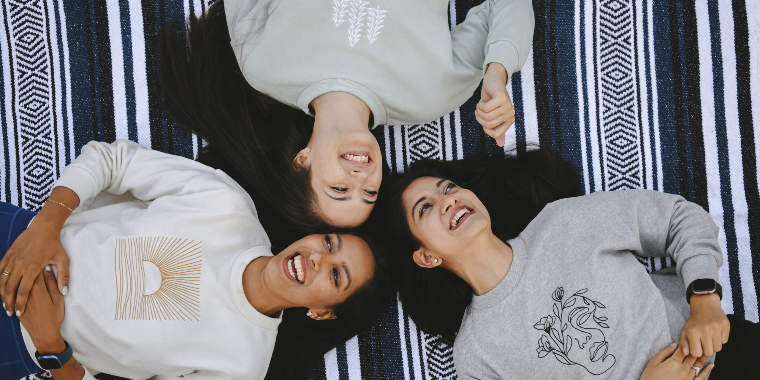 Three people smiling laying on a picnic blanket wearing sustainably made sweaters by Happy Earth.