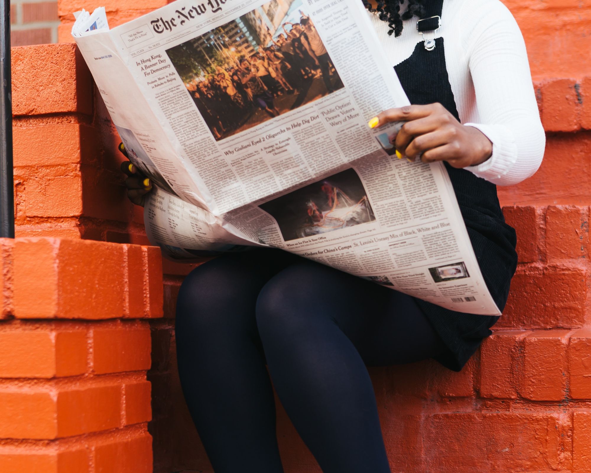 Someone in a black and white stylish outfit sitting on a red brick stoop and reading a newspaper.