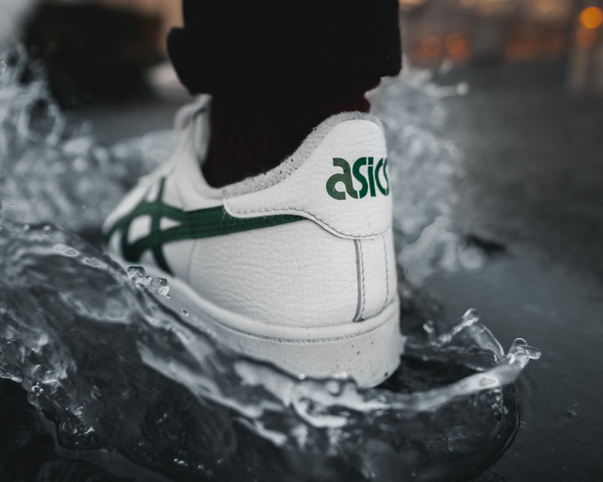 A pair of white and green Asics sneakers splashing into a puddle.