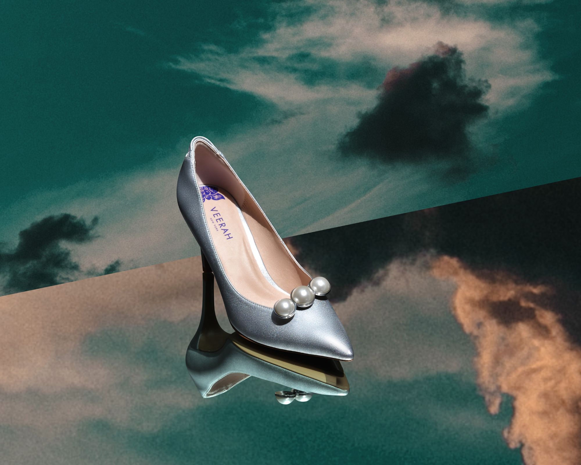 A pair of silver vegan VEERAH pumps with a trinity brooch on the front, with a moody cloudy background.